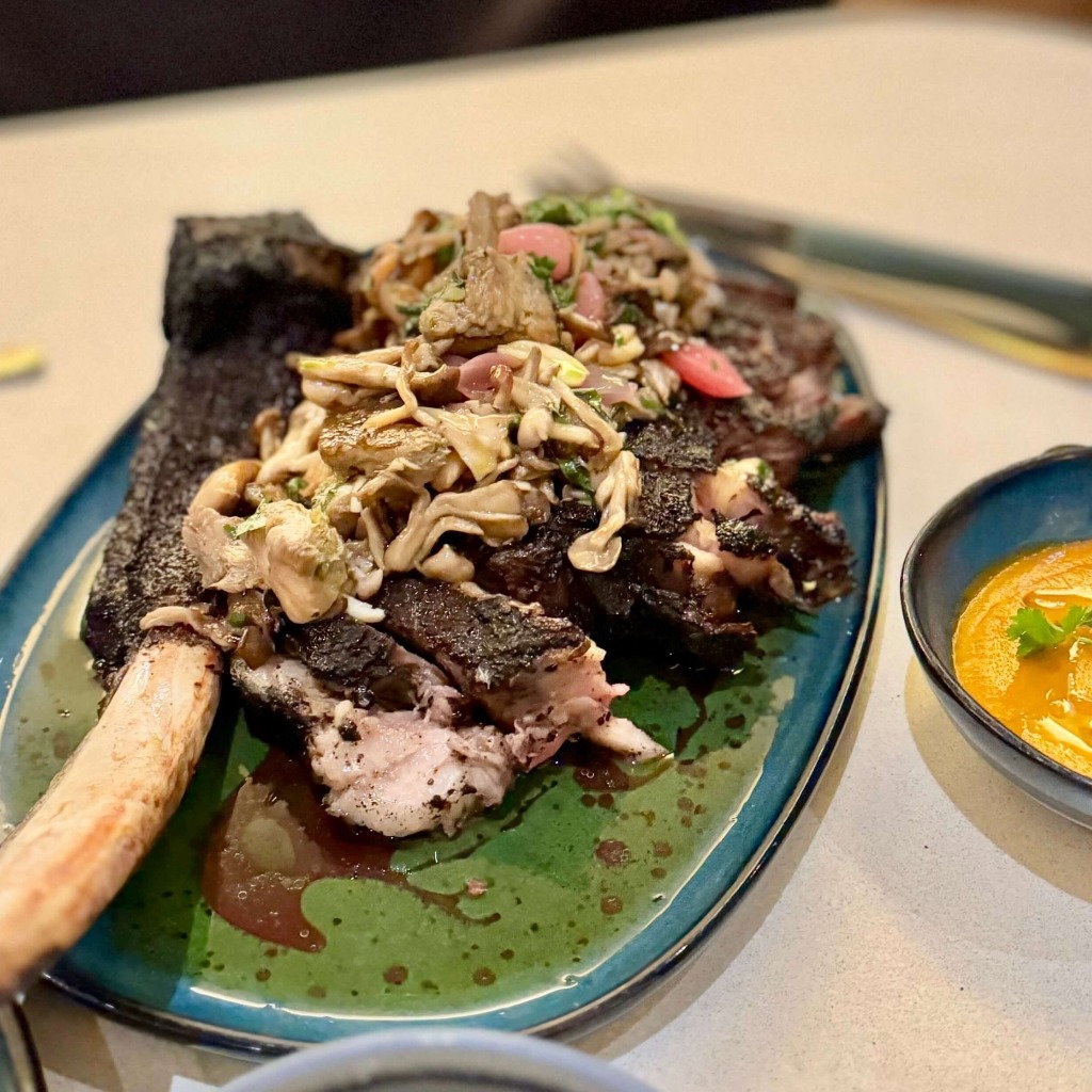 Grilled rib with a topping of fresh mushrooms and herbs, served on a ceramic plate accompanied by a bowl of smooth, creamy orange sauce garnished with cilantro leaves.