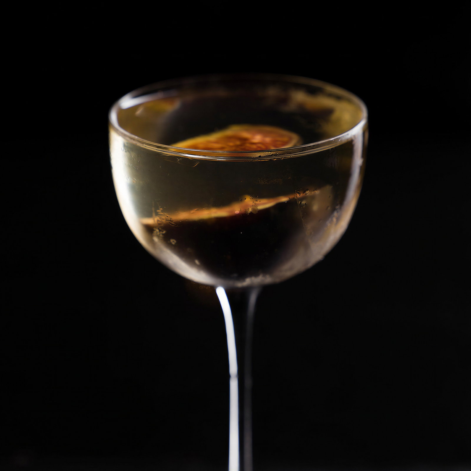 A cocktail glass with a dark liquid and an orange slice, set against a black background, creating an elegant and moody atmosphere.