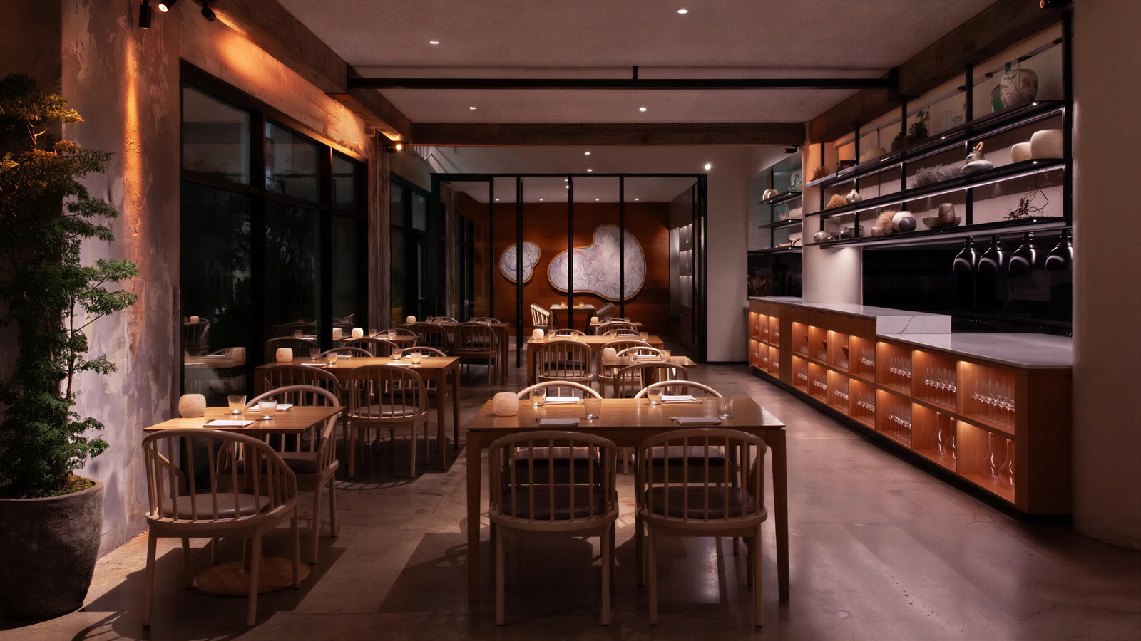Empty, elegant restaurant interior with wooden tables and chairs, warm lighting, abstract wall art, and glassware on shelves. Calm ambiance.