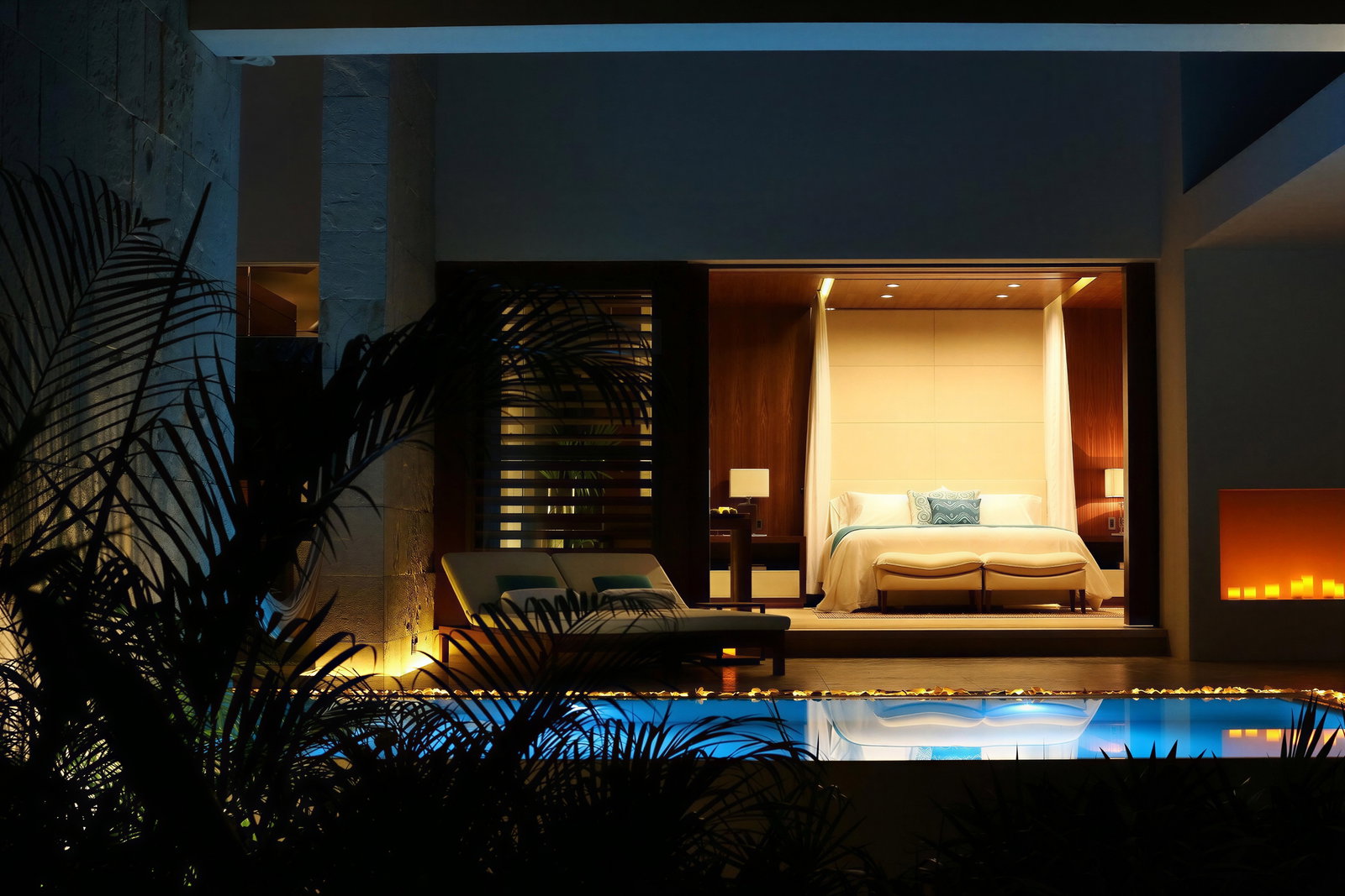 Luxurious bedroom with a lit pool in the foreground. Warm tones, modern decor, palm shadows, and a cozy ambiance create a serene mood.