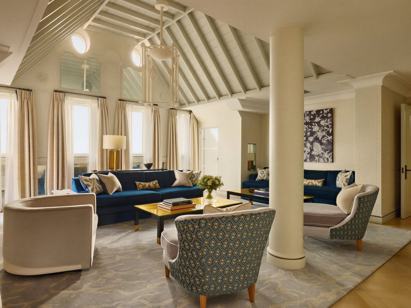 Spacious living room with blue sofas, patterned chairs, and large windows. Beige and blue decor. A pillar and floral artwork are visible.