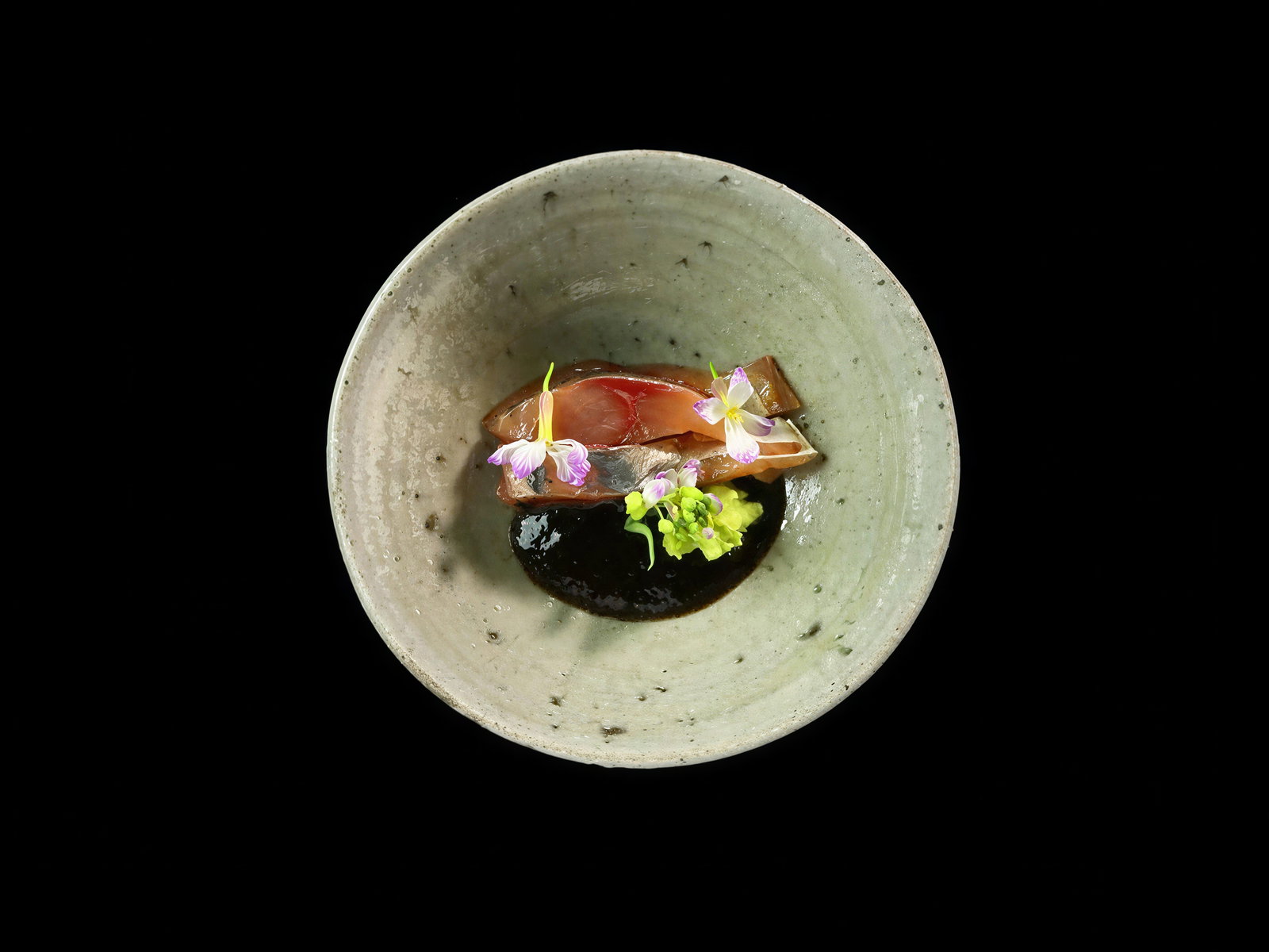 A ceramic bowl on a black background holds a gourmet dish with garnishes and vibrant flowers, creating an artistic and elegant presentation.