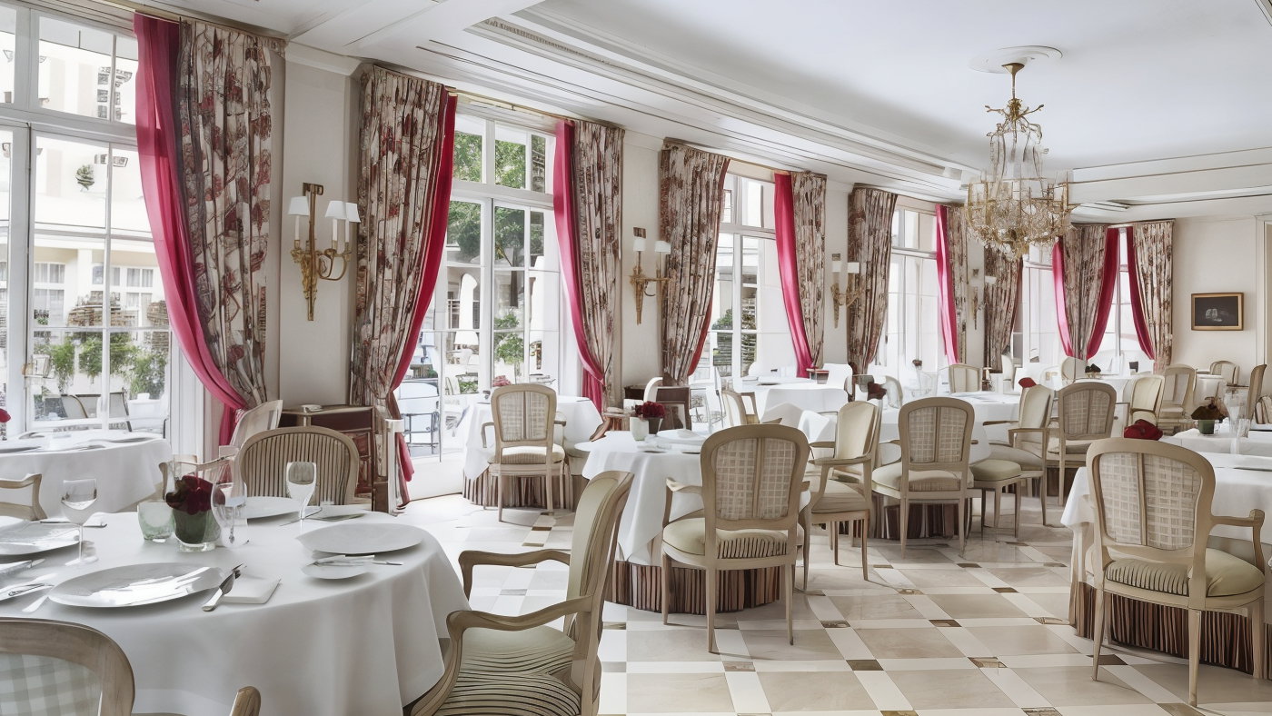 Elegant restaurant with white tablecloths, beige chairs, and red-accented curtains. Chandeliers hang overhead, creating a formal ambiance.