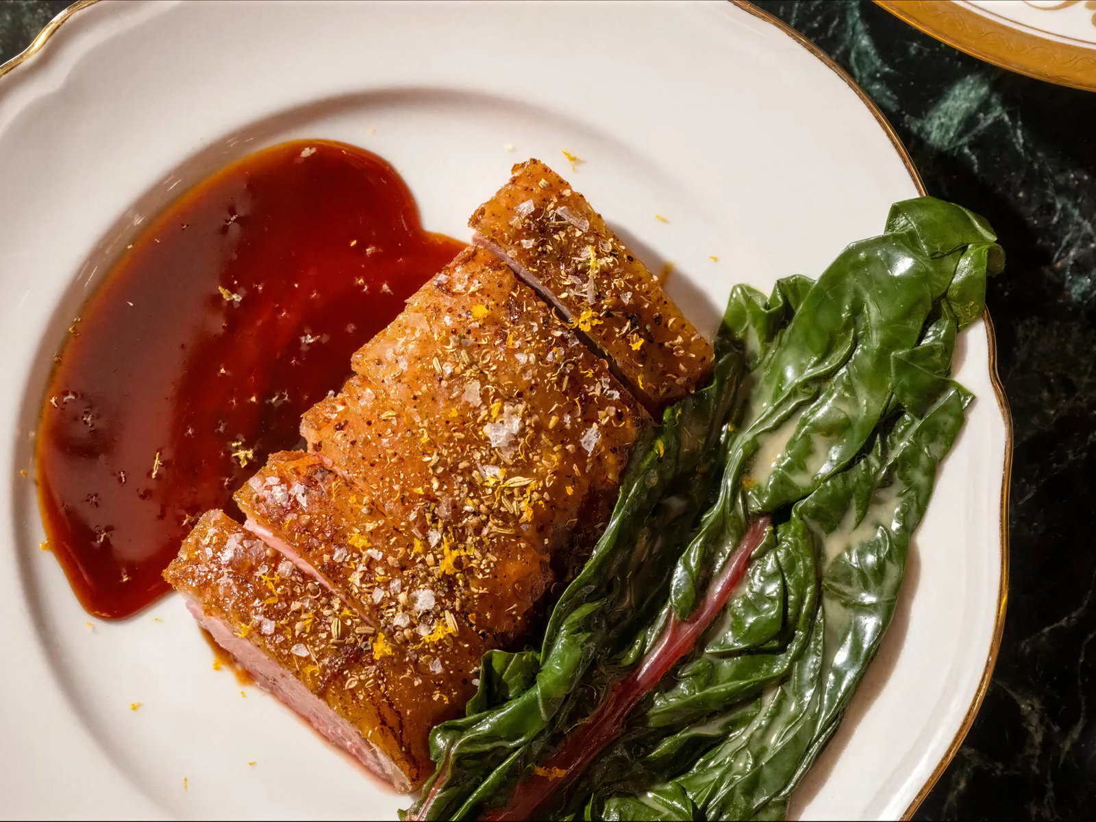 Crispy sliced pork with glaze and greens on a white plate against a dark marble background, featuring golden spices and a rich brown sauce.