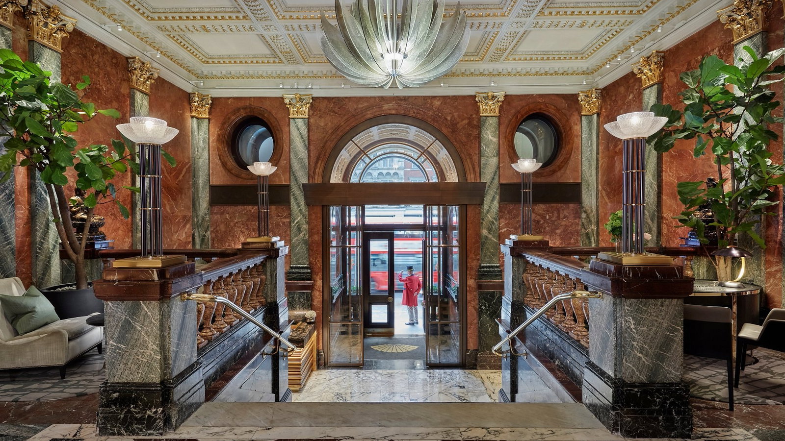 Elegant hotel lobby with marble, gold accents, and plants. A doorman in a red coat holds the door open against a backdrop of passing traffic.