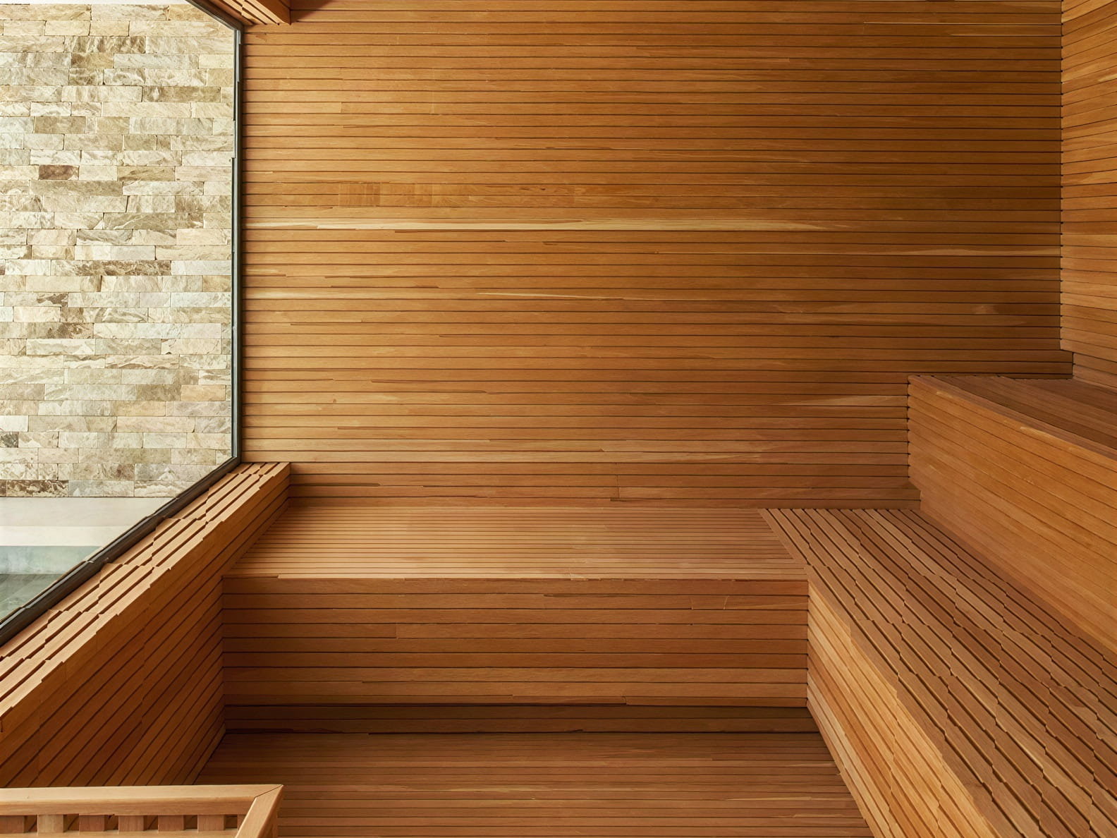 Wooden sauna interior at Hotel Humano Spa with tiered seating and a stone wall. Natural light streams through a glass window, creating a warm, serene atmosphere.