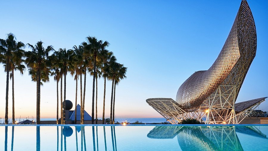 Sunset view of a reflective pool, tall palm trees, and a large golden fish sculpture in a modern architectural setting, creating a serene ambiance.