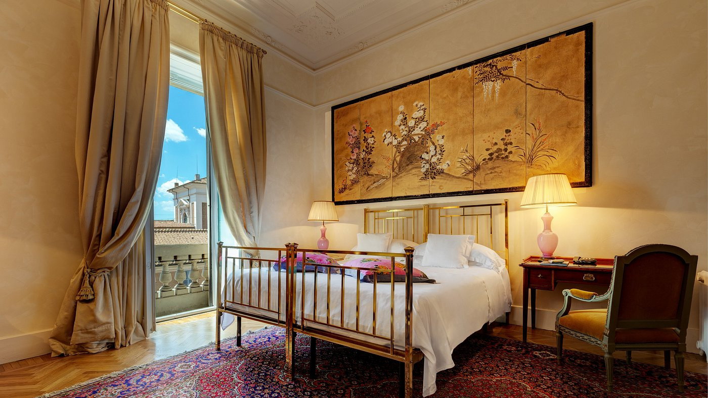 Elegant bedroom with a large bed, golden headboard, and floral artwork. Warm lighting from lamps, plush rug, and a view through open curtains.