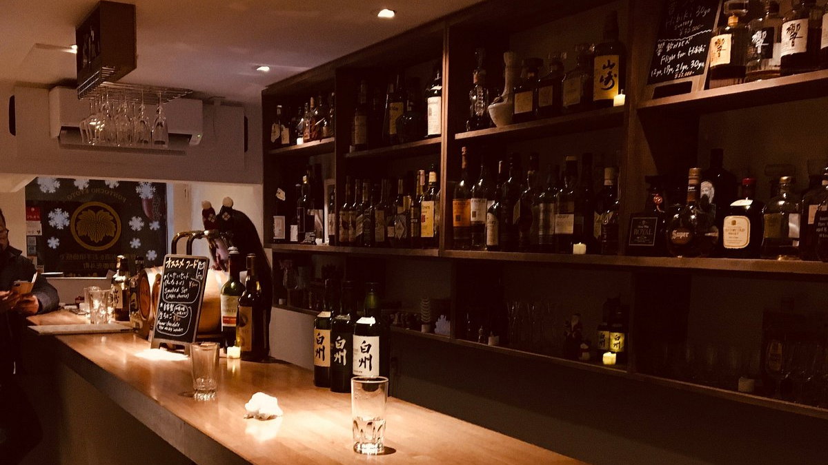 Dimly lit bar with shelves of assorted liquor bottles, chalkboard menu, and a wooden countertop. Two people sit quietly, creating a cozy ambiance.