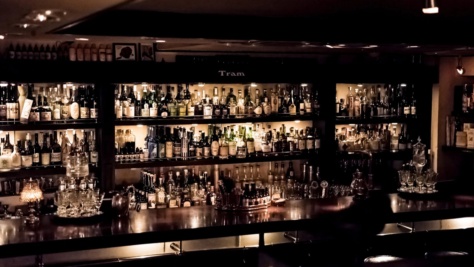 Dimly lit bar interior with shelves full of assorted liquor bottles. A wooden counter with glasses and a soft, warm glow creates a cozy ambiance.