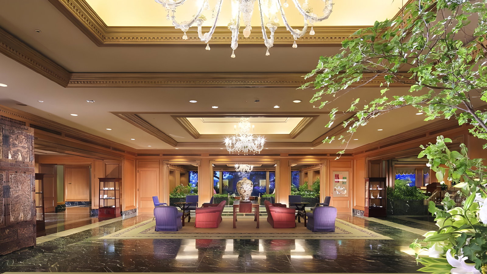Elegant hotel lobby with chandeliers, red and purple chairs, marble floor, and lush green plants, creating a luxurious and welcoming atmosphere.