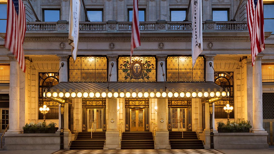 Elegant hotel entrance with warm lights, flags above, and glass doors. Checkerboard floor and ornate details create a grand ambiance.