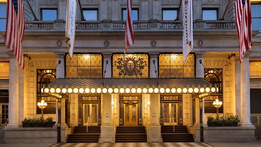 Elegant hotel entrance with warm lights, flags above, and glass doors. Checkerboard floor and ornate details create a grand ambiance.