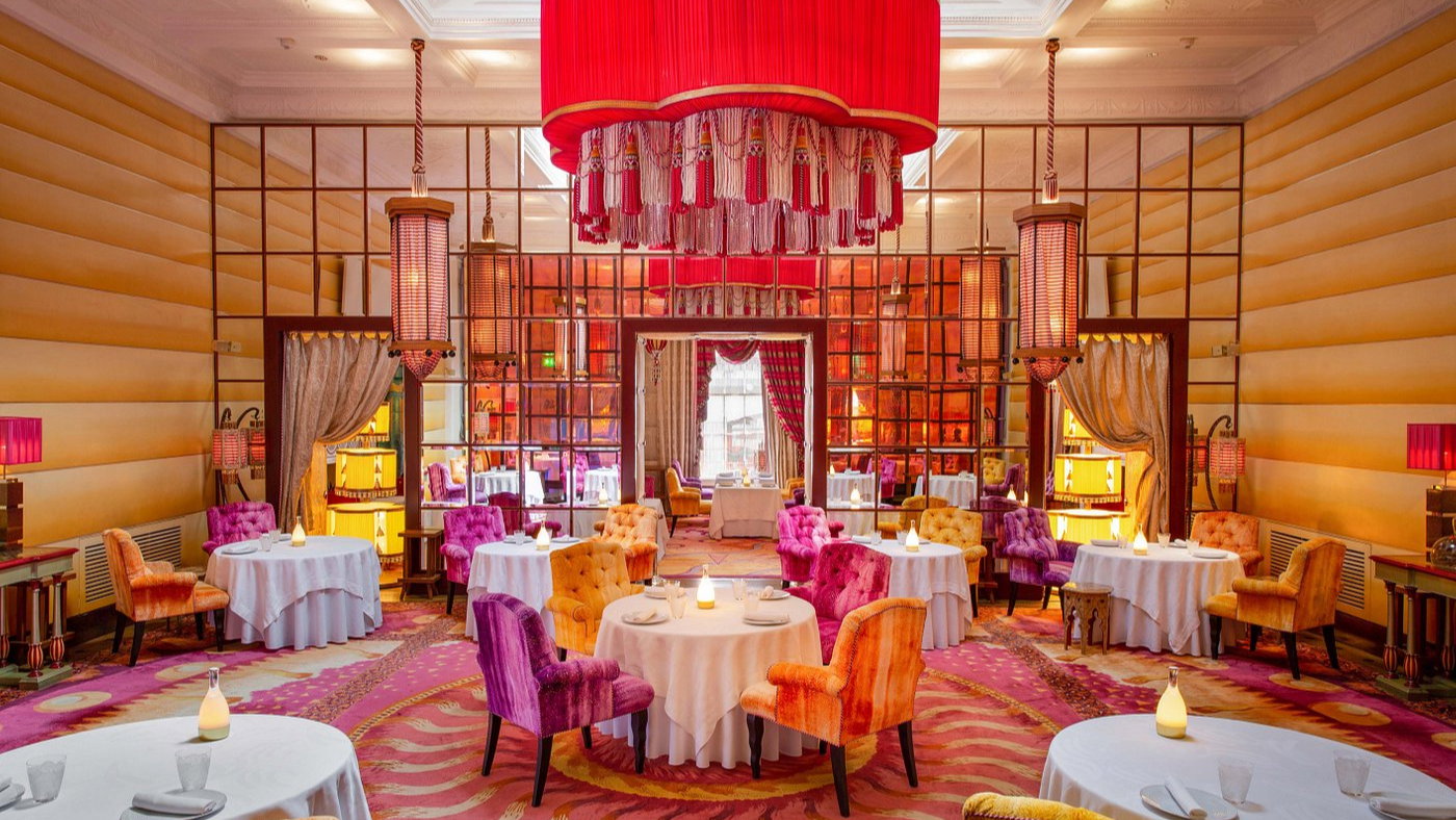 Elegant restaurant with vibrant orange and purple chairs, round tables, and red chandeliers. Warm lighting creates a cozy, luxurious ambiance.