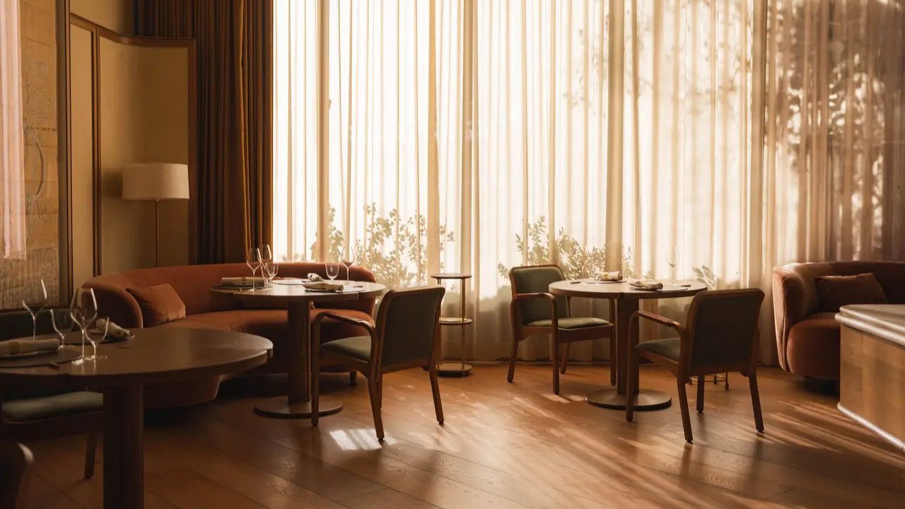 Cozy restaurant interior with round tables, chairs, and soft lighting. Floor-to-ceiling curtains diffuse sunlight, creating a warm atmosphere.