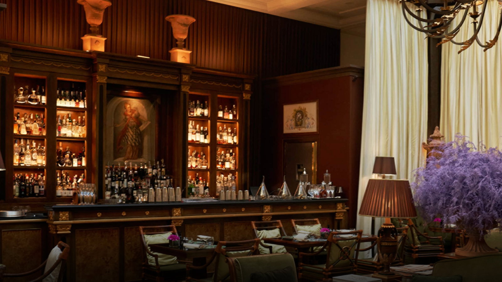 Elegant bar with bottles on wooden shelves, ornate columns, a large purple floral arrangement, warm lighting, and tall cream curtains.