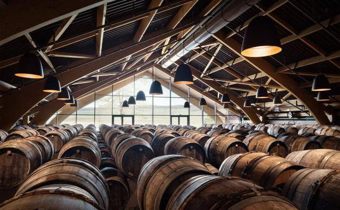 Wooden barrels line a rustic warehouse with high ceilings and large windows. Ambient lighting and symmetrical beams create a tranquil mood.