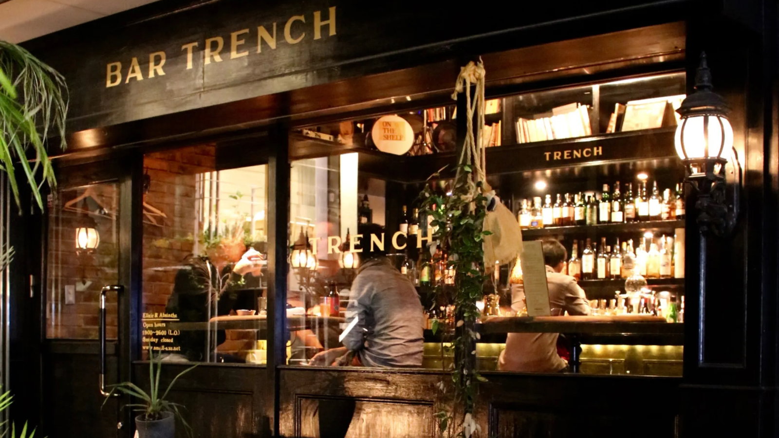 People inside Bar Trench in a cozy setting, seated at the bar with bottles on shelves. A menu is visible. Warm lighting and plants enhance ambiance.