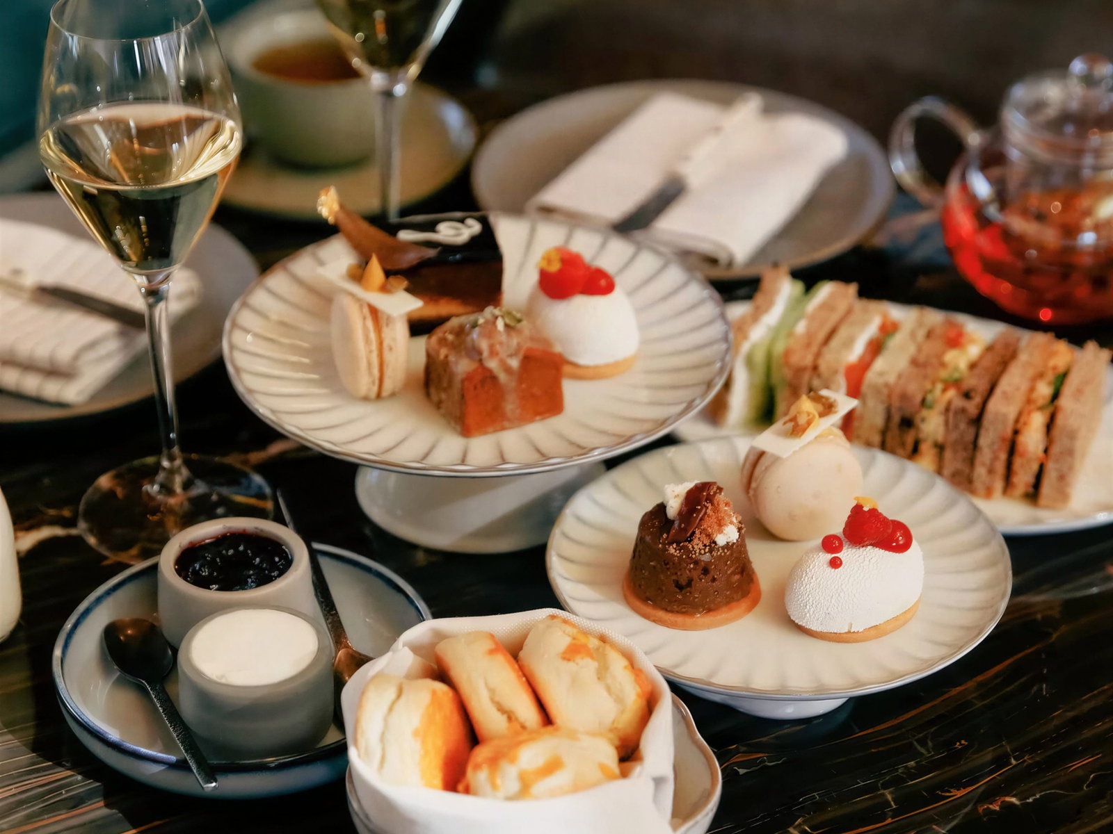 Elegant afternoon tea setting with assorted pastries, sandwiches, and scones on plates. Wine and tea cups are visible. Cozy atmosphere.