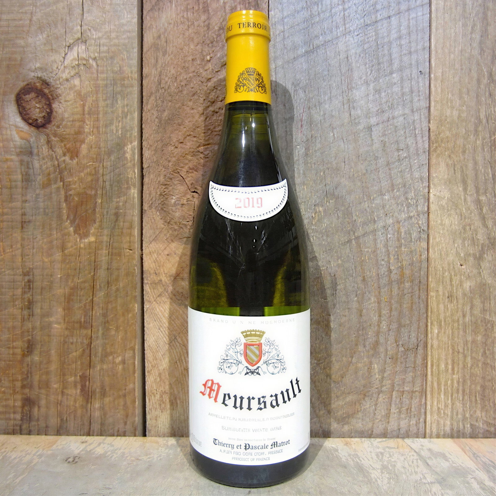 A bottle of Meursault wine with a yellow cap, labeled 2019, stands on a wooden surface with a textured wood background.