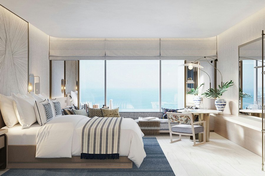 Elegant hotel room with large window overlooking the ocean. Neutral tones, cozy bed with striped blanket, floral chair, and potted plants.