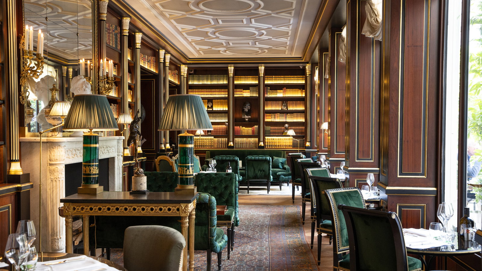 Elegant library lounge with green velvet chairs, ornate lamps, and bookshelves. Warm lighting, classic wood paneling, and a cozy atmosphere.