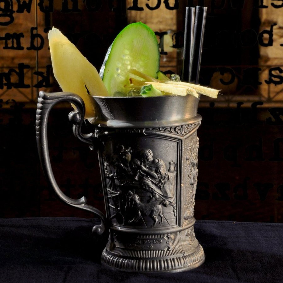 Ornate metal mug with lime, cucumber slices, and two straws. Background features reversed black text on glass. Vintage, elegant mood.