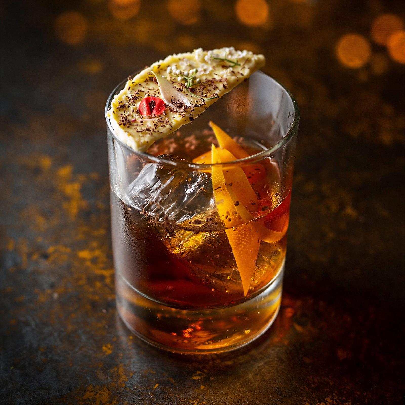 A cocktail in a glass with ice and orange peel, garnished with a decorative slice. Warm, moody lighting and a dark, textured background.