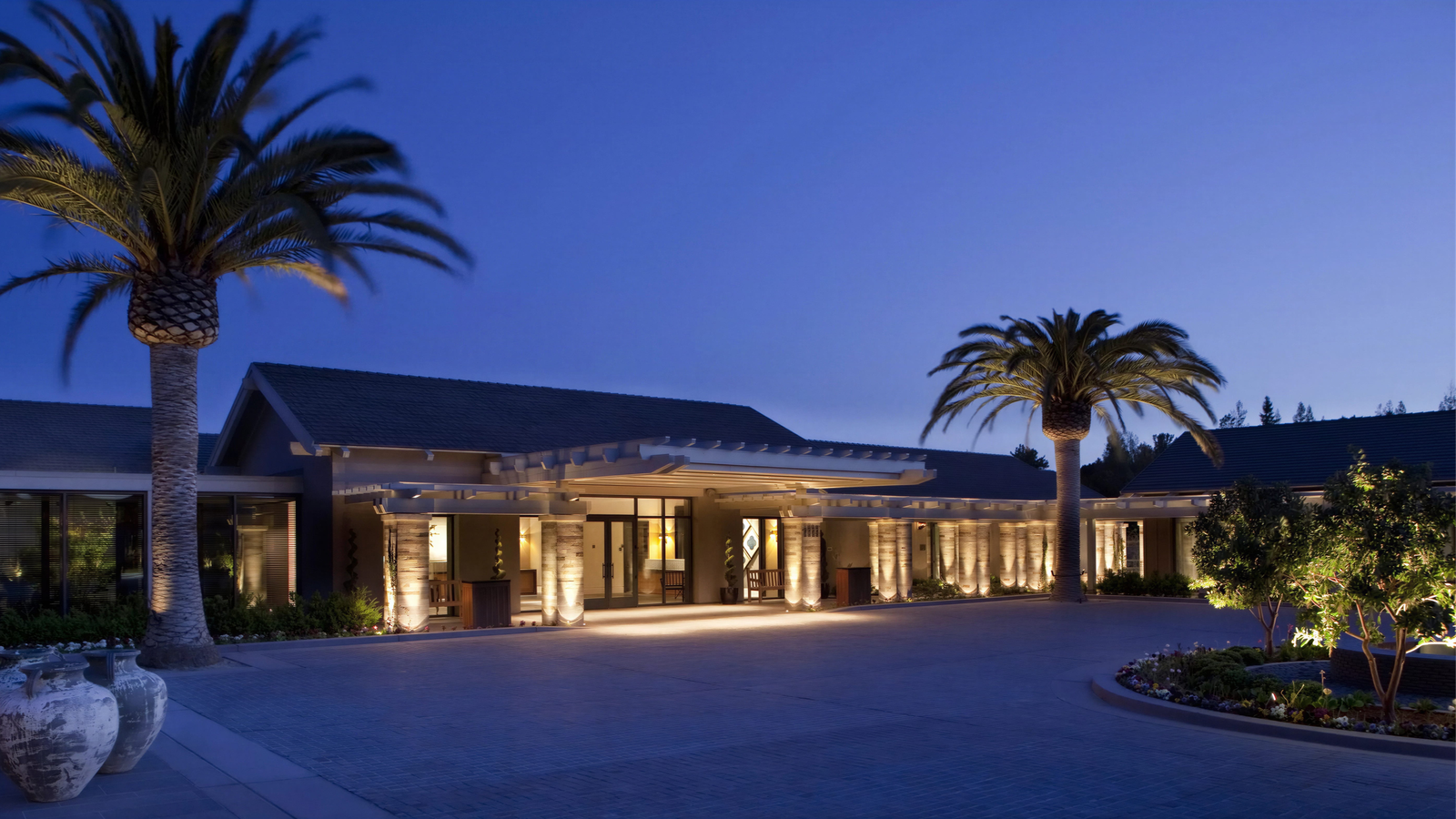 Modern building with palm trees, warmly lit at twilight. Stone columns and potted plants enhance the serene evening atmosphere.
