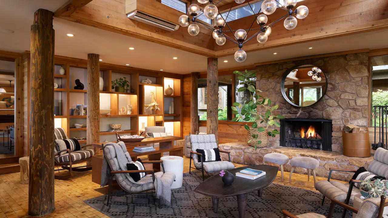 Cozy living room with stone fireplace, modern chandelier, and cushioned chairs. Wooden shelves hold decor. Warm lighting and rustic charm.