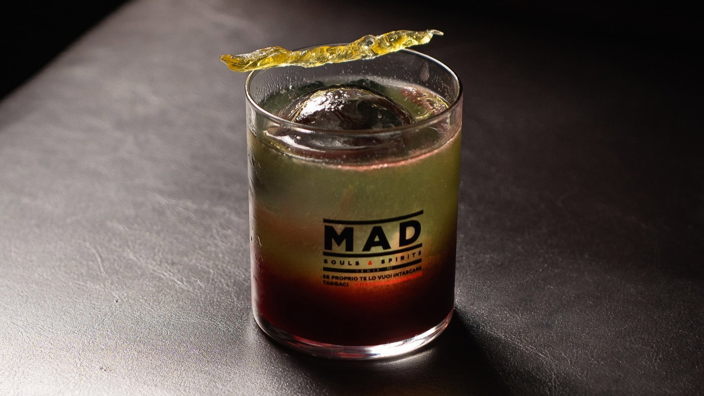 A cocktail with gradient colors, topped with a thin garnish, sits on a dark surface. The glass has "MAD" text. Elegant and refined mood.