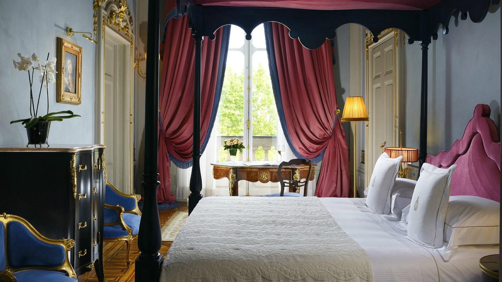 Luxurious bedroom with ornate four-poster bed, pink drapes, blue and gold chairs, and elegant decor. Bright, serene ambiance.