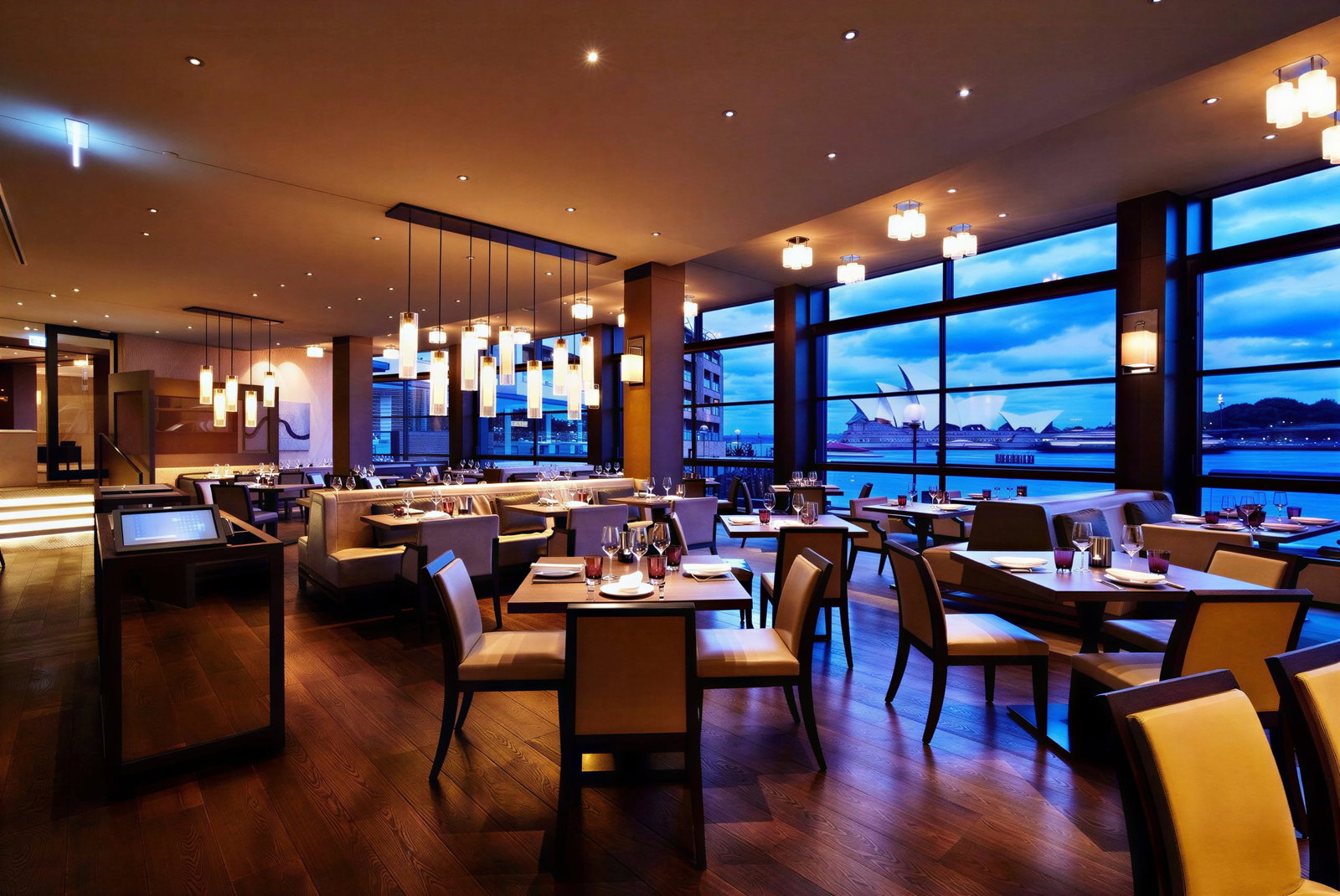 The Dining Room by James Viles at Park Hyatt Sydney interior with modern lighting, wooden floors, and set tables. Large windows reveal a view of a city skyline at dusk.