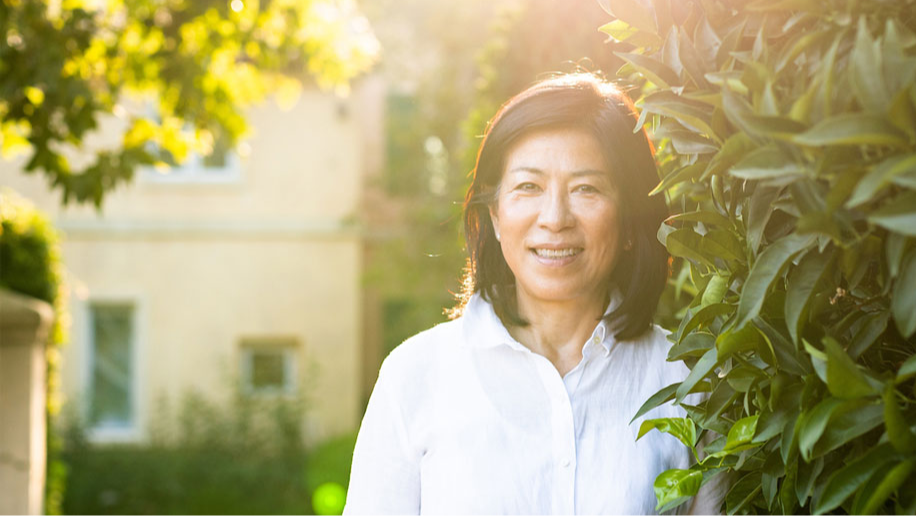 Smiling woman in white shirt stands beside green bushes, with sunlit yellow house in background, creating a warm, cheerful atmosphere.