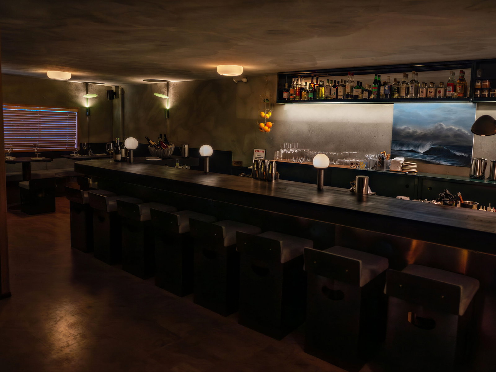 Dimly lit bar and dining room of The dining room at Smithereens New York with dark wooden counter, lined with empty stools. Bottles sit on shelves against a seascape painting, creating a cozy mood.