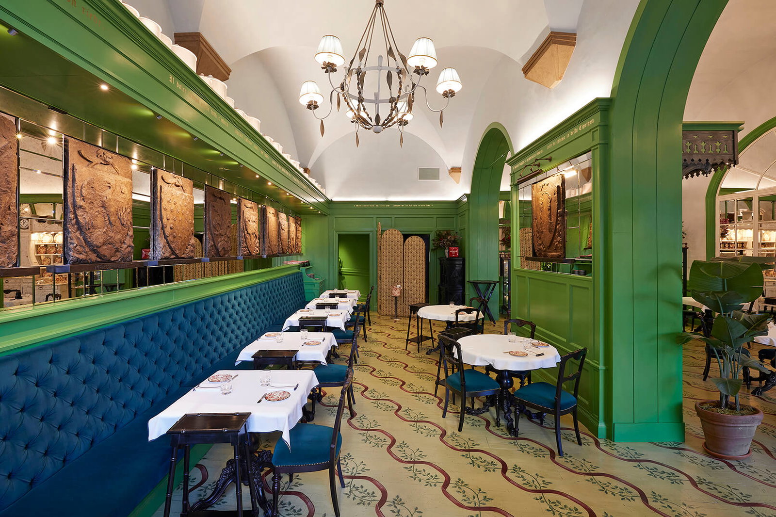 Elegant restaurant with green walls, ornate lighting, blue seating, and white tablecloths. Art and plants adorn the space, creating a cozy vibe.