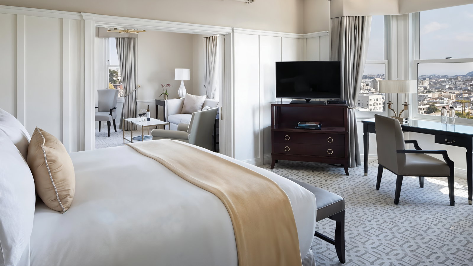 Elegant hotel room with a cozy bed, a beige throw, and pillows. A TV stands opposite. Large window reveals city view. Sophisticated decor.