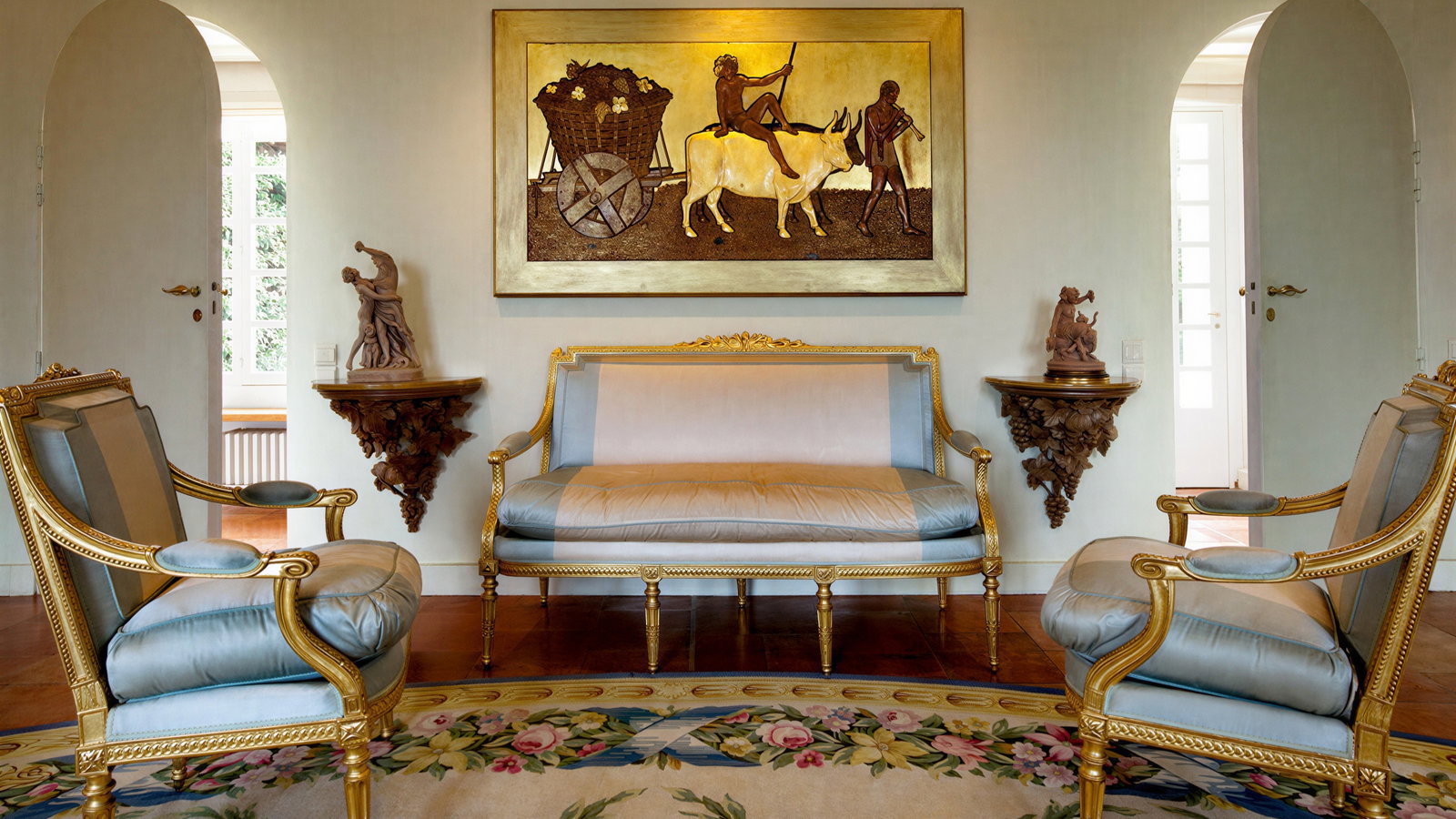 Elegant room with blue and gold furniture, two sculptures, and a painting of figures with an ox. Ornate floral rug and arched doorways.