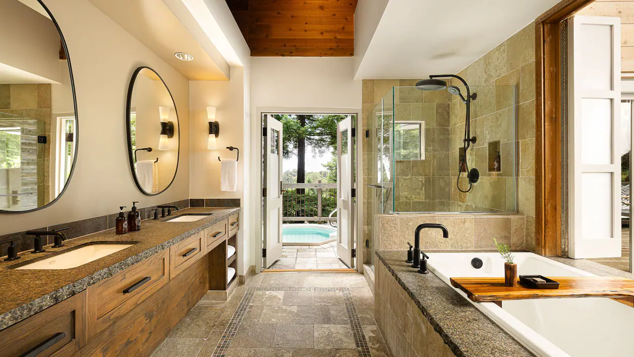 Elegant bathroom with dual sinks, large mirrors, and a glass shower. Opens to a patio with a pool, surrounded by greenery.