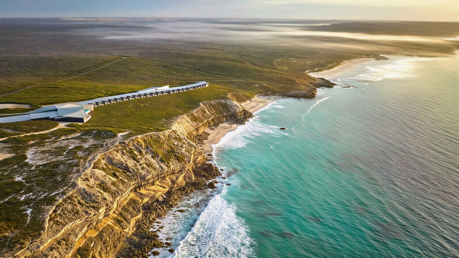 Aerial view of cliffside Southern Ocean Lodge which borders a vast coastline, with turquoise waves crashing against rugged rocks. The lush green landscape stretches inland.