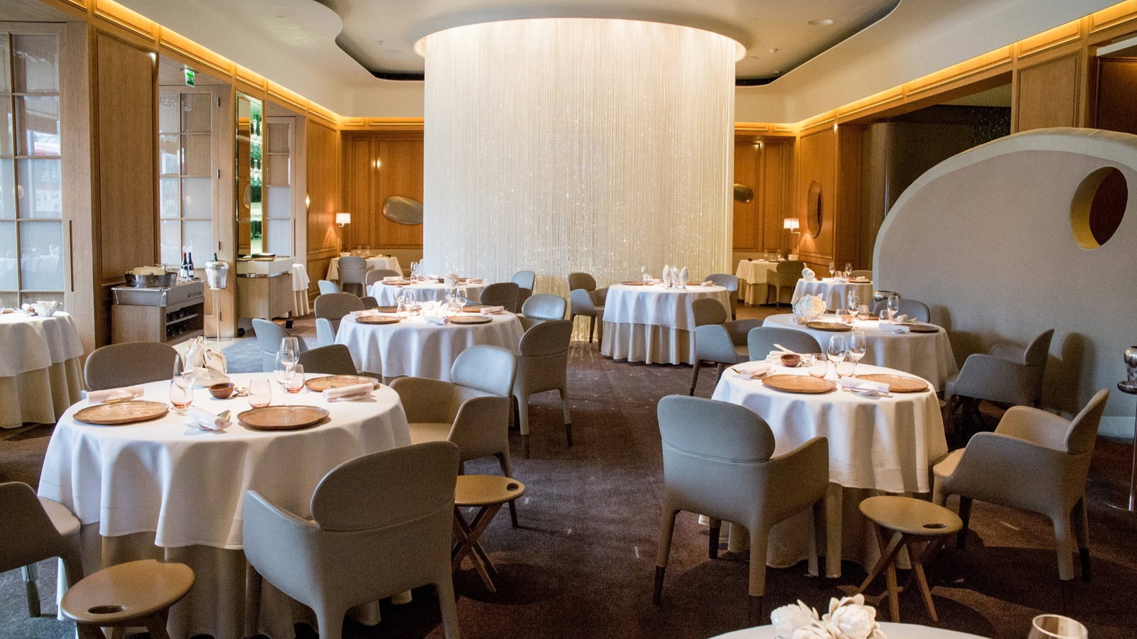Elegant restaurant interior with round tables, white tablecloths, and beige chairs. A glowing cylindrical light fixture dominates the center.