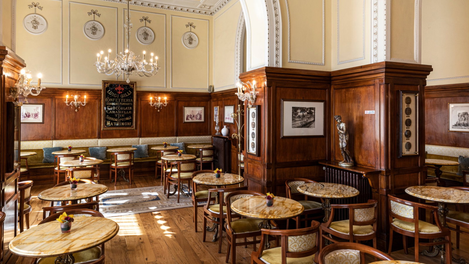 Elegant café interior with marble tables, wooden paneling, chandeliers, and framed art. A vintage sign reads "Luigi Gilli Confetteria."