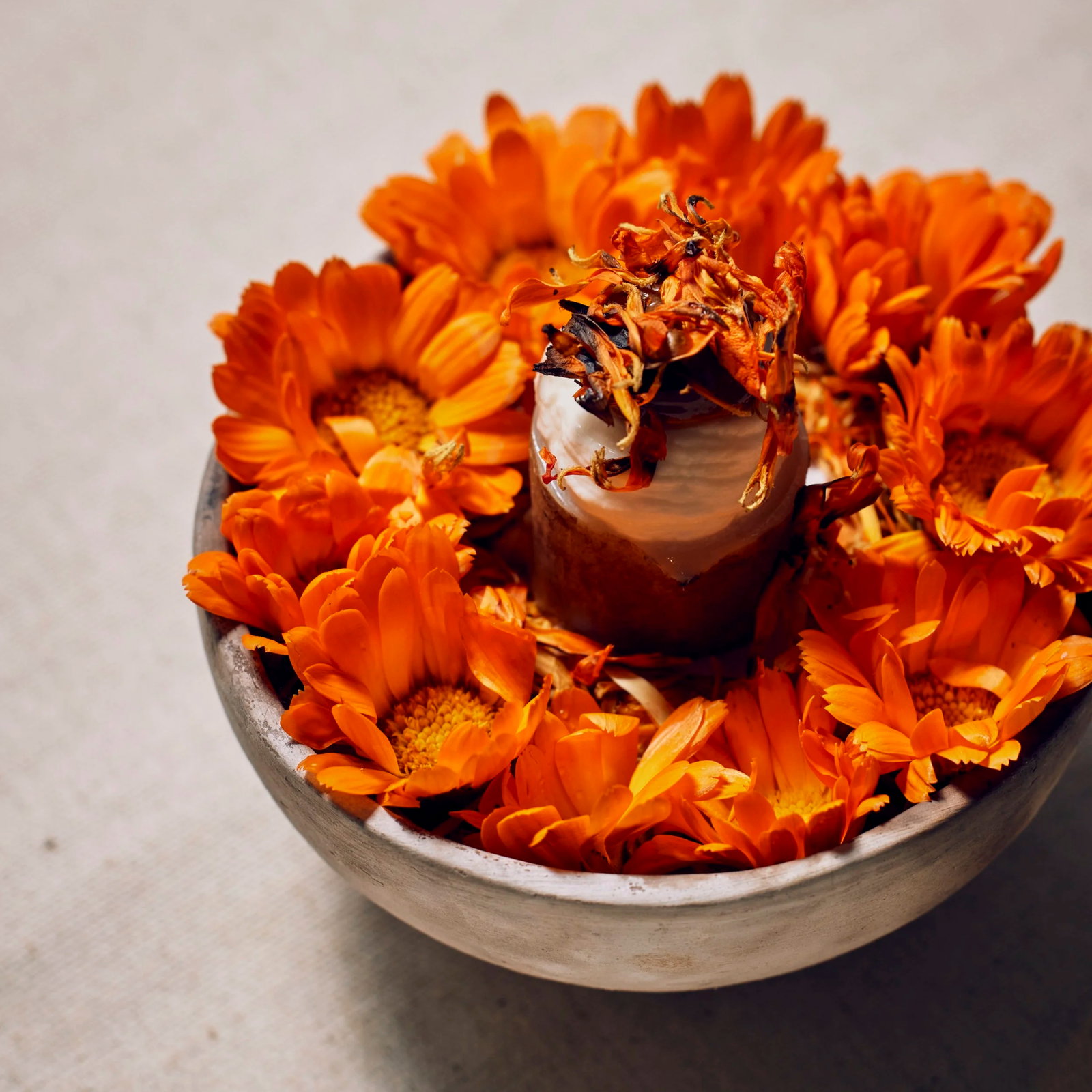 A ceramic bowl filled with vibrant orange marigold flowers surrounds a dessert topped with dried petals, set on a textured light surface.