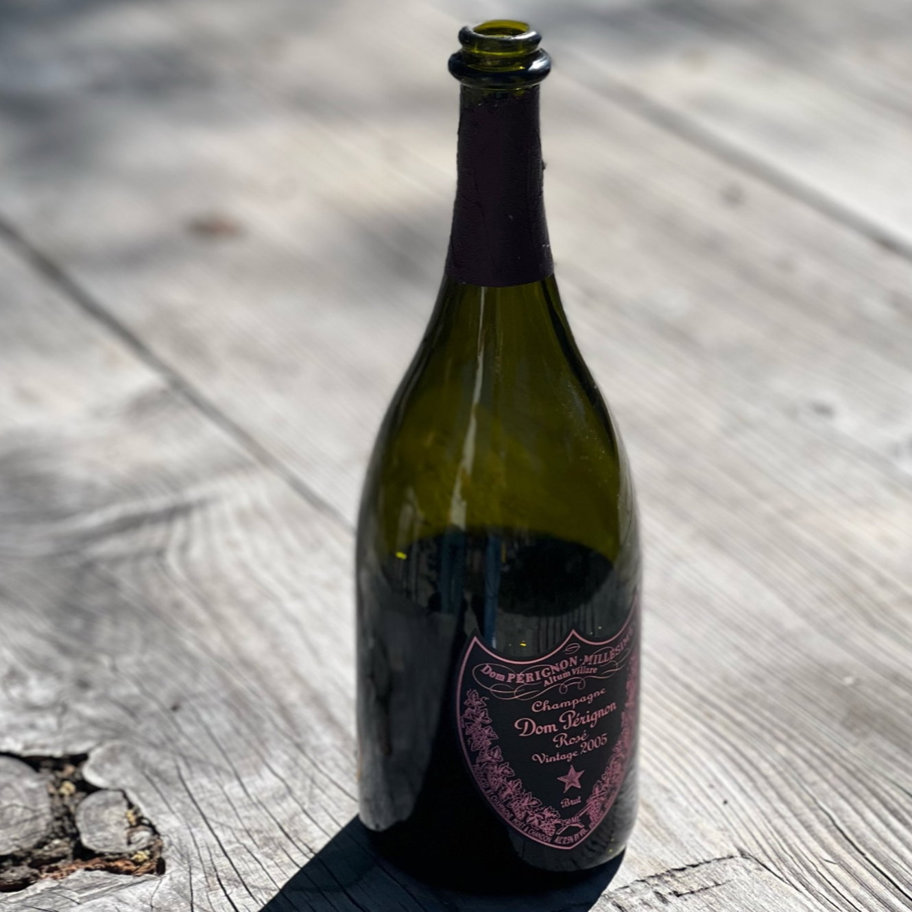 Empty Dom Pérignon champagne bottle on sunlit wooden table. Pink label reads "Vintage 2005." Shadow cast across planks.