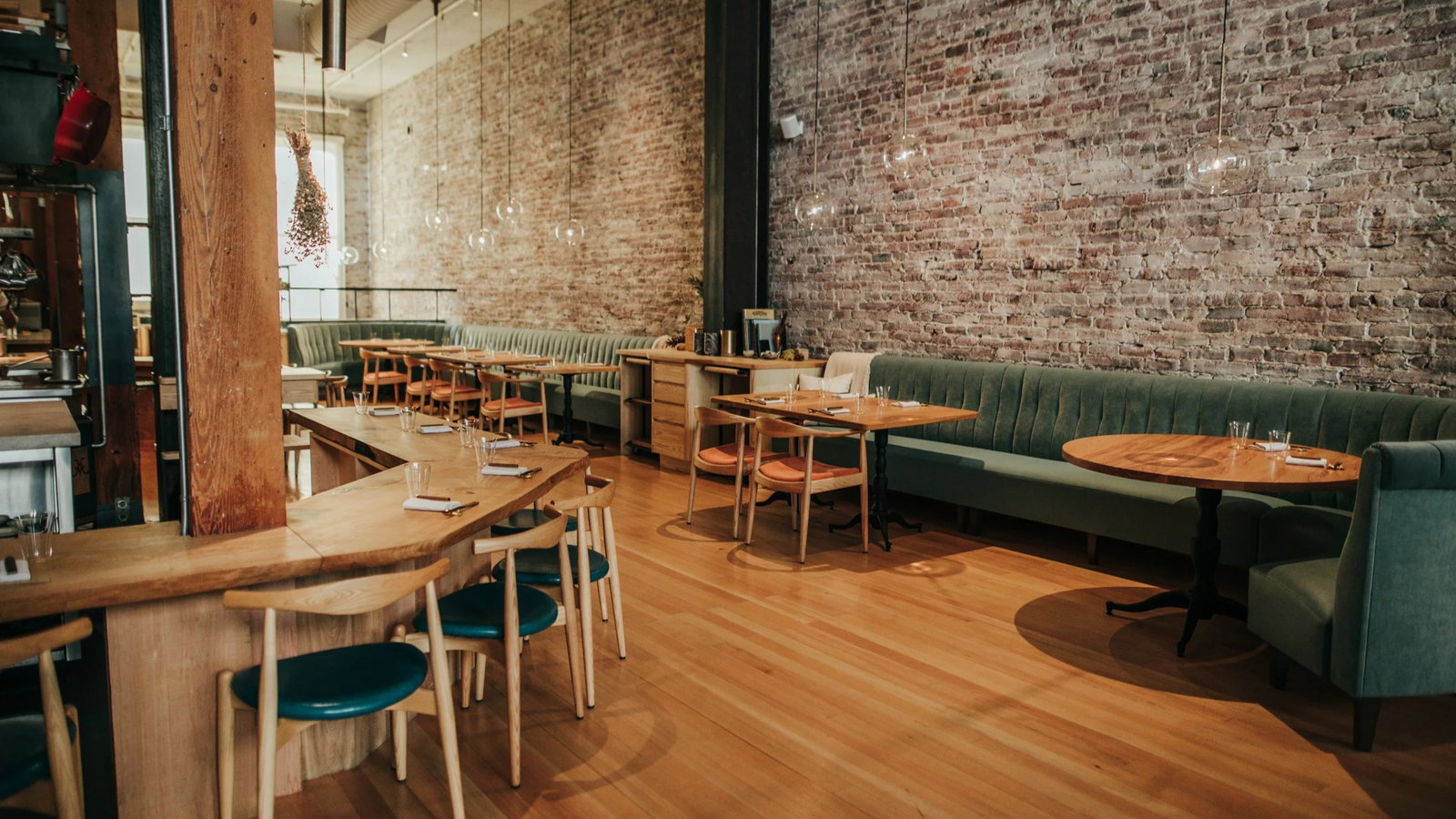 Cozy restaurant interior with wooden tables, teal chairs, and green booths against exposed brick walls. Warm lighting, inviting atmosphere.