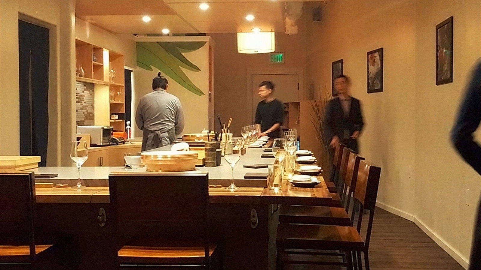 Restaurant interior with wooden tables set for dining, a chef in a gray robe prepares food, two people walk by. Warm lighting, artwork on walls.