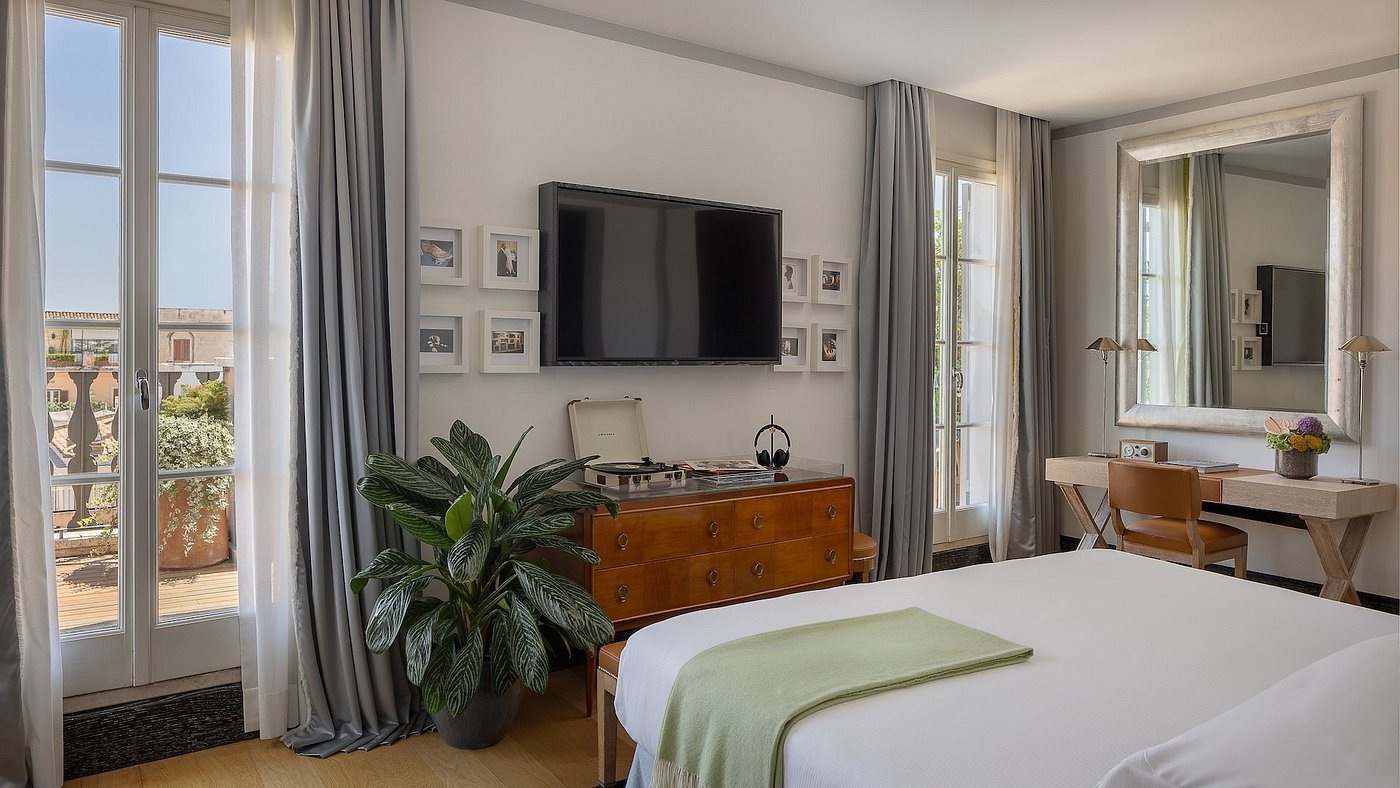 Bright bedroom with a large bed, TV, and a wooden dresser topped with a record player and plant. Open balcony doors with a city view.