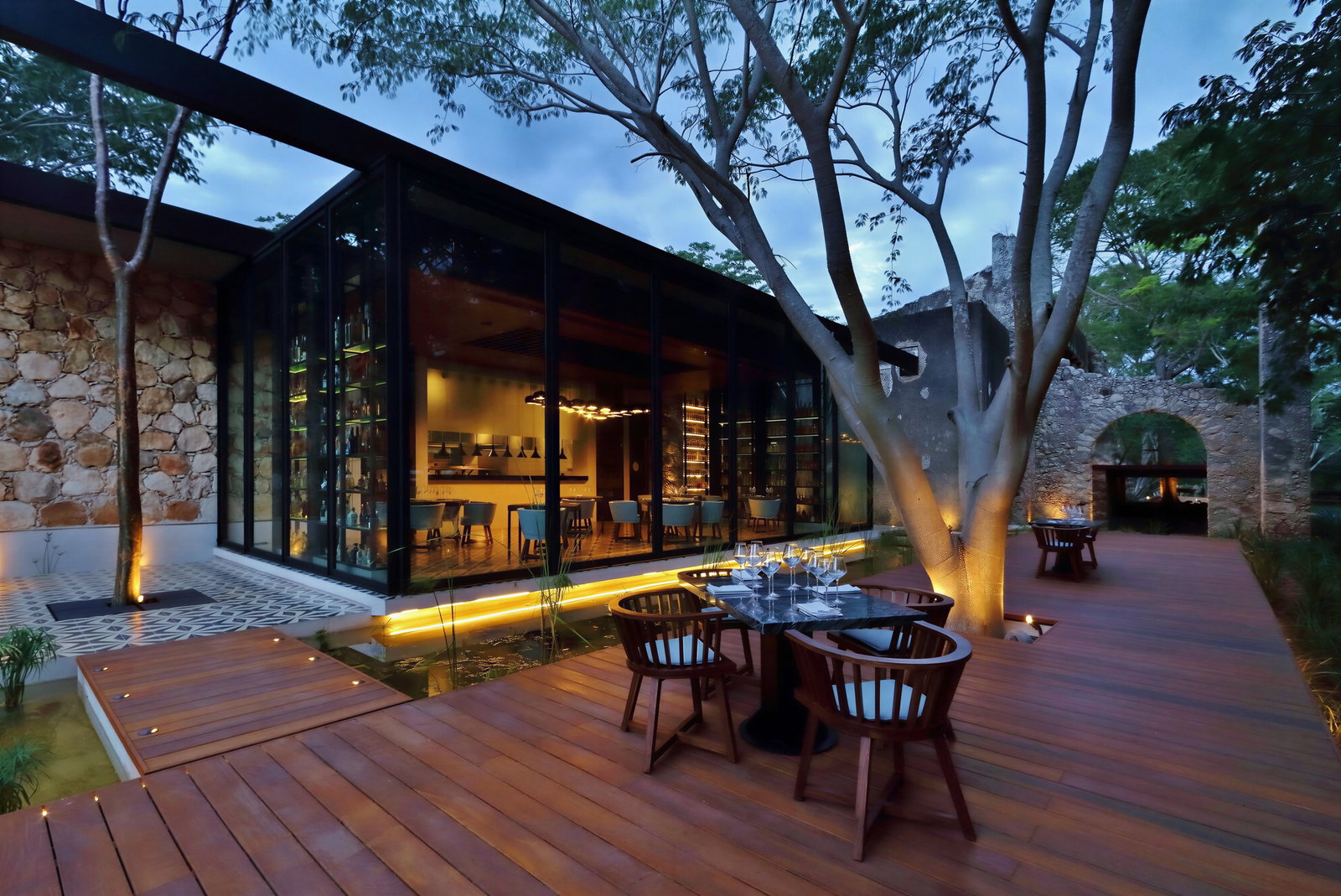 Modern restaurant exterior with large glass windows, warm lighting, and wooden deck. Empty tables with chairs, surrounded by trees at dusk.