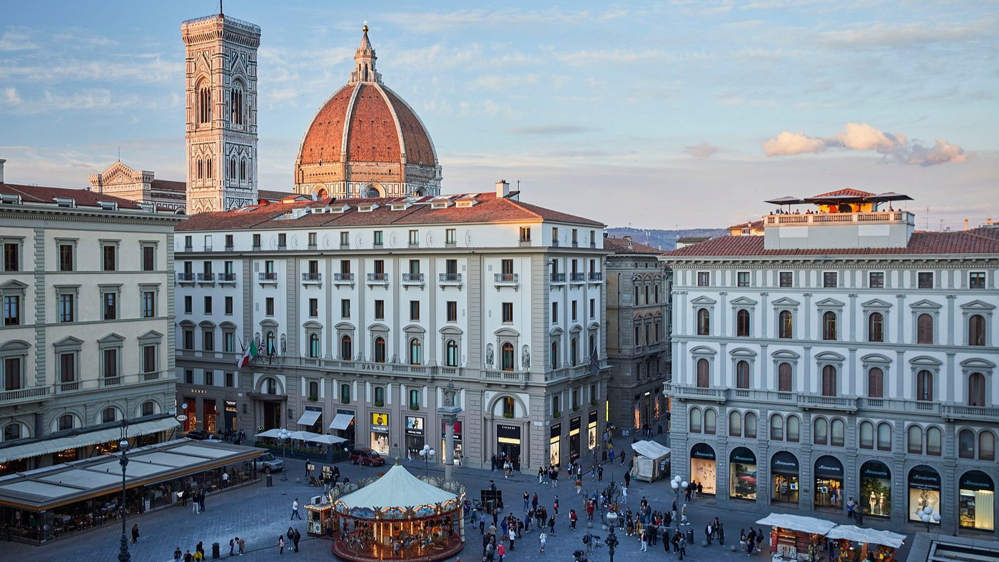 A busy square in Florence with people walking, a carousel, and historic buildings. The Duomo and bell tower stand tall under a blue sky.