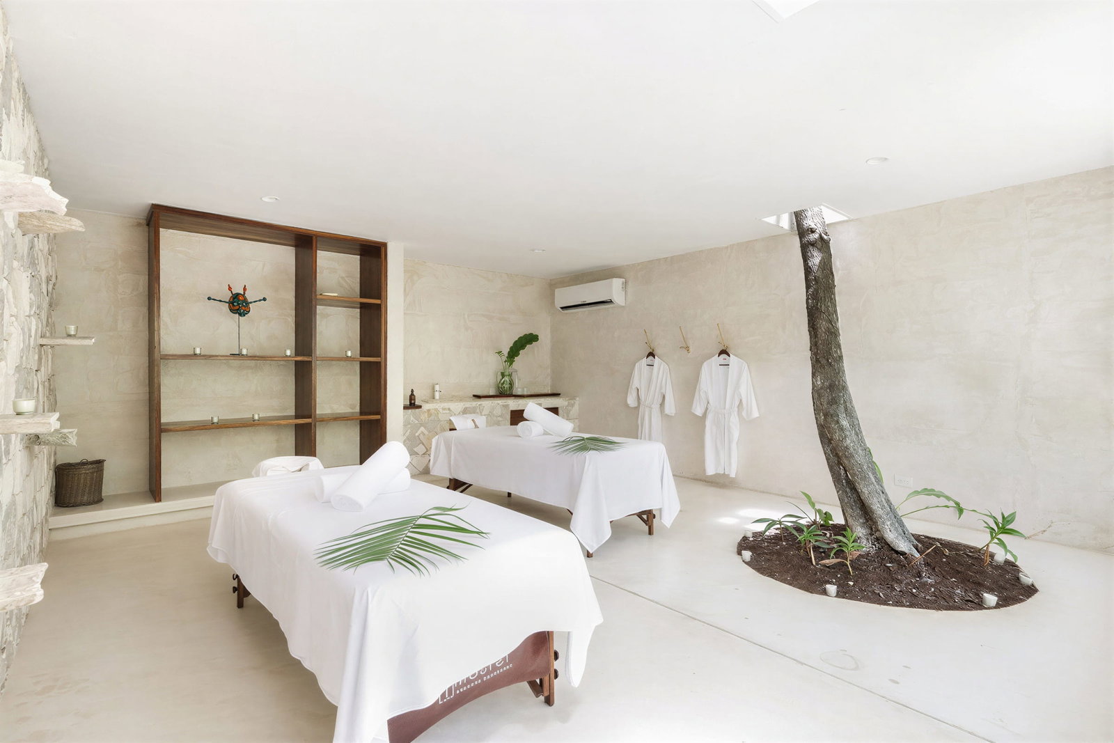 Massage room at Wakax Hacienda with two tables covered in white sheets and leaves. Stone wall with shelves, robes hanging, and a tree indoors. Calm ambiance.